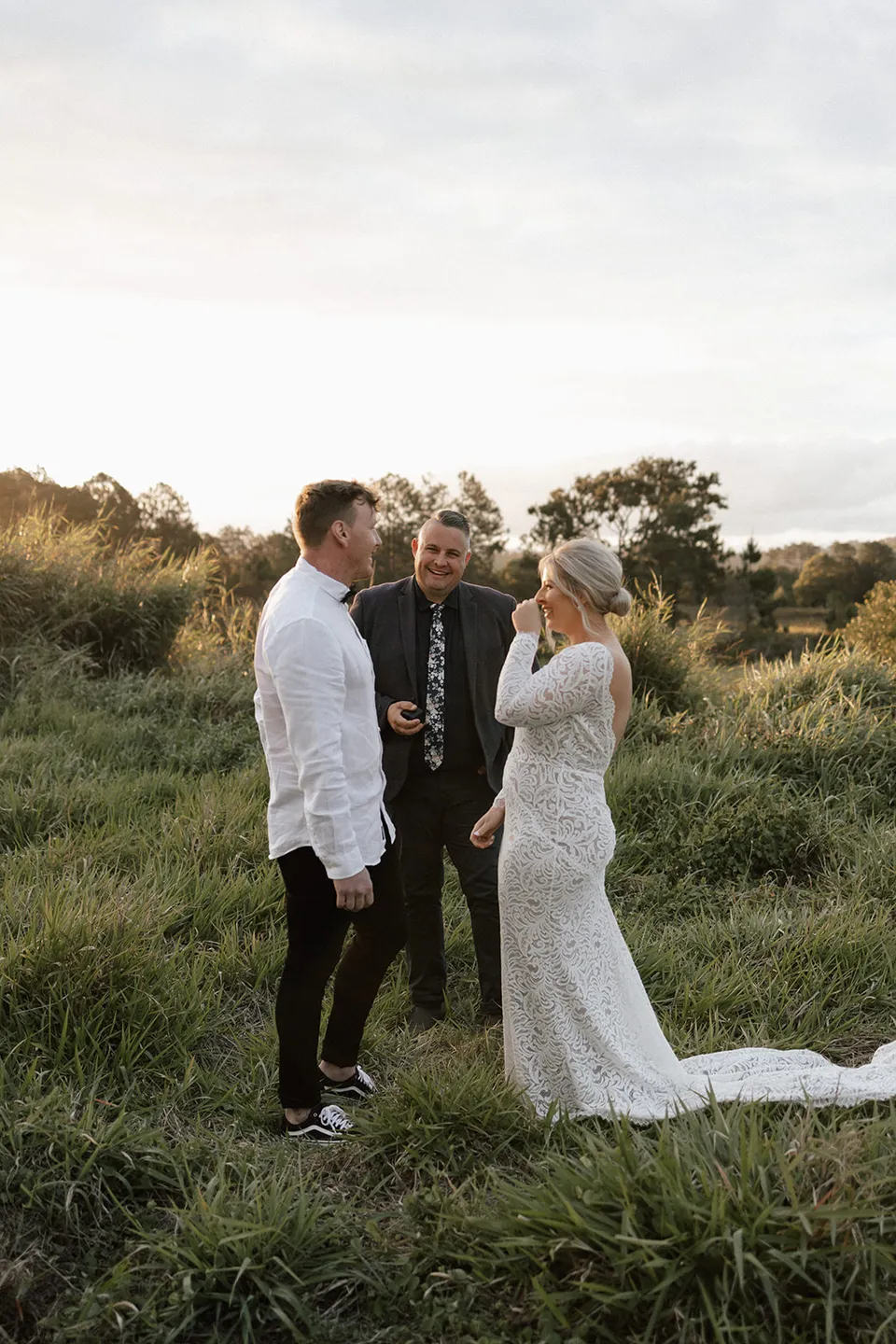 Laura raising a hand to her eye as Matt and celebrant Josh Withers stand with her in the sunlit grass, in the Tweed Valley, NSW, for an Elopement Collective elopement. Photo by Bulb Creative.