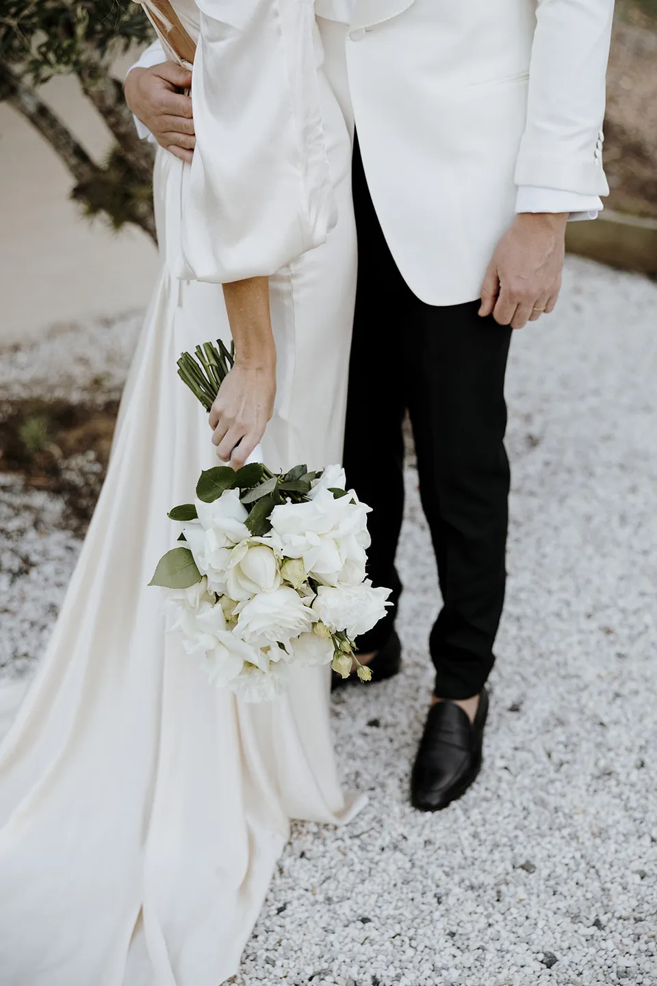 Close view of Gabrielle’s white bouquet and draped ivory dress beside Greg’s white jacket and black trousers at Roundhill Retreat in Obi Obi, Sunshine Coast Hinterland, for their Elopement Collective elopement.