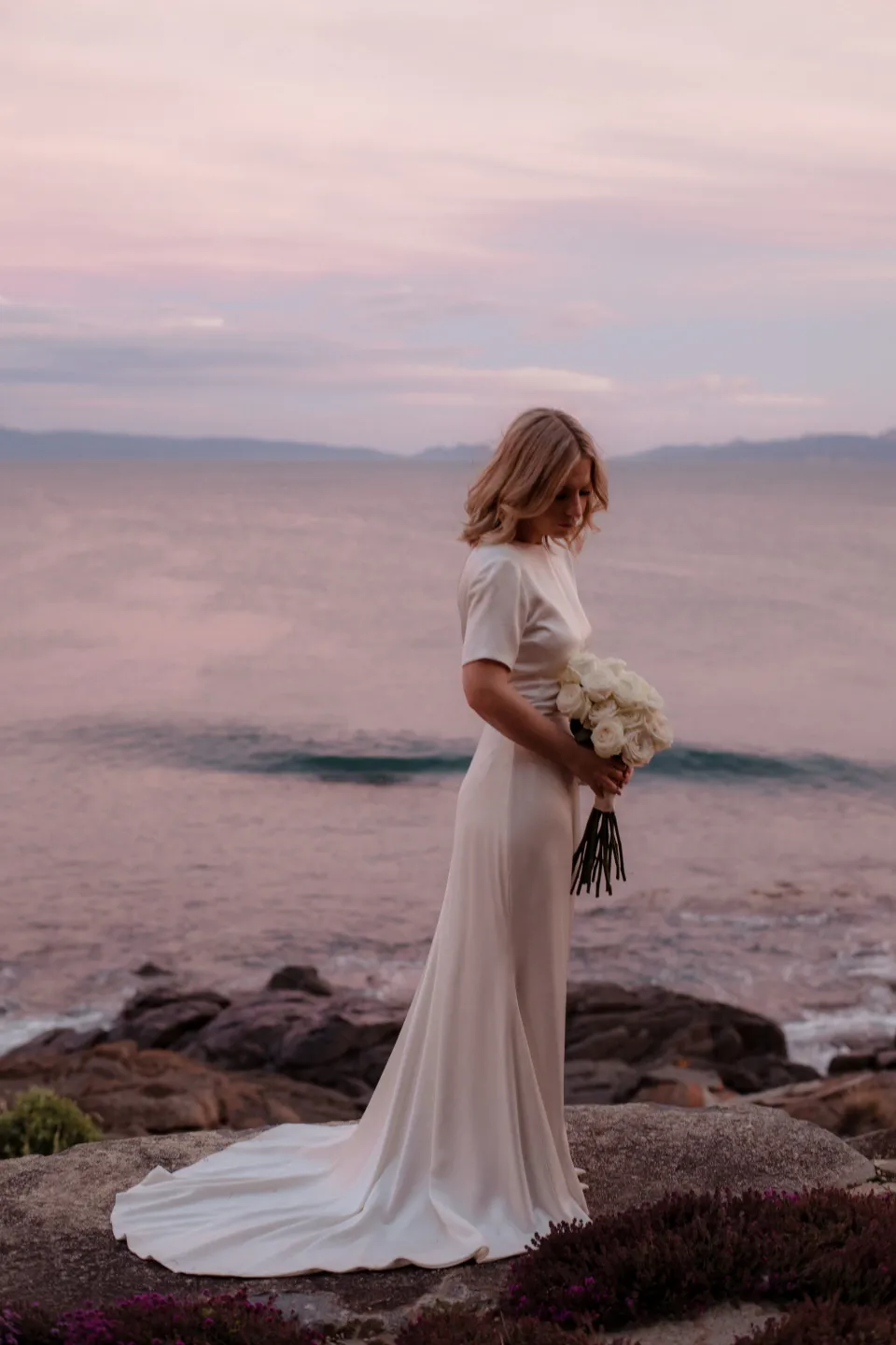 Jemmah holding her bouquet by The Romantics beside the bay beneath a pink sky, at Thalia Haven on the East Coast of Tasmania, Australia, for an Elopement Collective elopement. Photograph by Joey Willis.