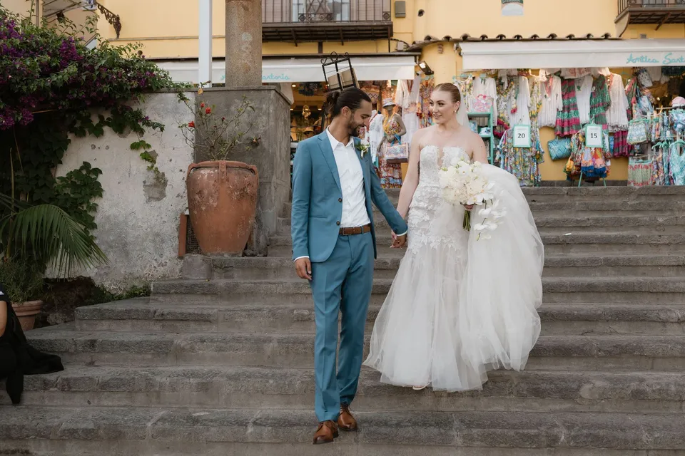 Skyler and Zach holding hands as they walk down stone steps past colourful shop displays, carrying her Flora Garden - Positano bouquet, in Positano on the Amalfi Coast, Italy, for an Elopement Collective elopement. Photograph by Jason Corroto.