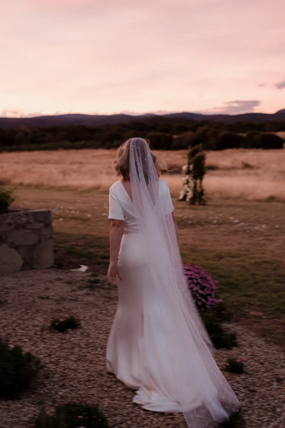 Rear view of Jemmah in her veil beneath a pink sunset sky, at Thalia Haven on the East Coast of Tasmania, Australia, for an Elopement Collective elopement. Photograph by Joey Willis.
