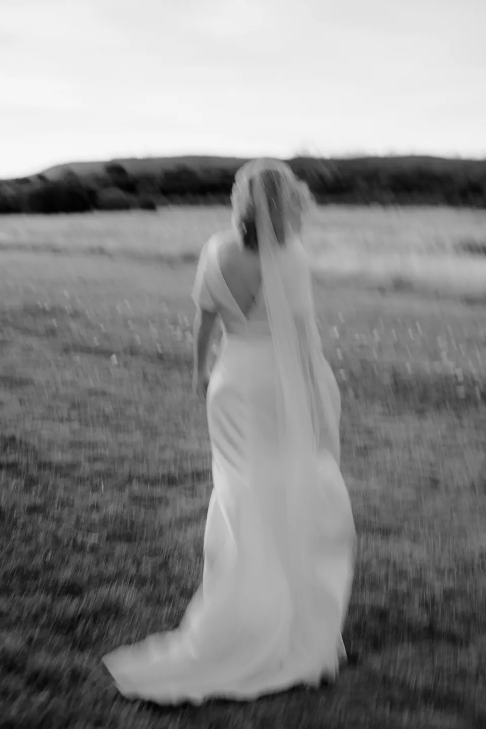 Black and white rear view of Jemmah standing among the meadow grasses, at Thalia Haven on the East Coast of Tasmania, Australia, for an Elopement Collective elopement. Photograph by Joey Willis.