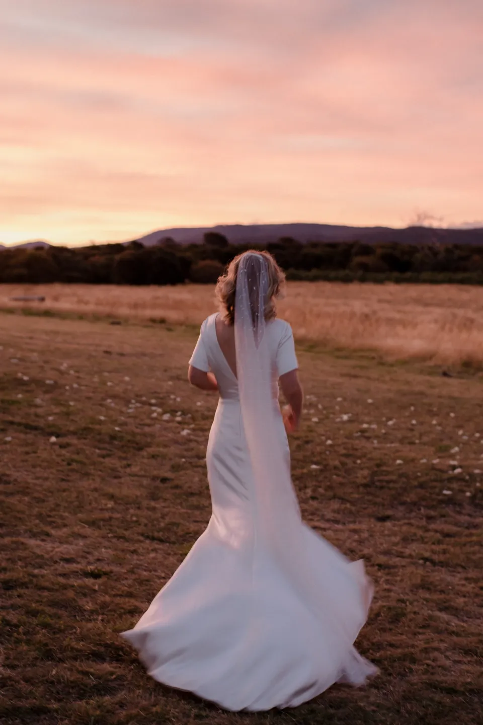 Jemmah looking across the meadow beneath a glowing pink sky, at Thalia Haven on the East Coast of Tasmania, Australia, for an Elopement Collective elopement. Photograph by Joey Willis.