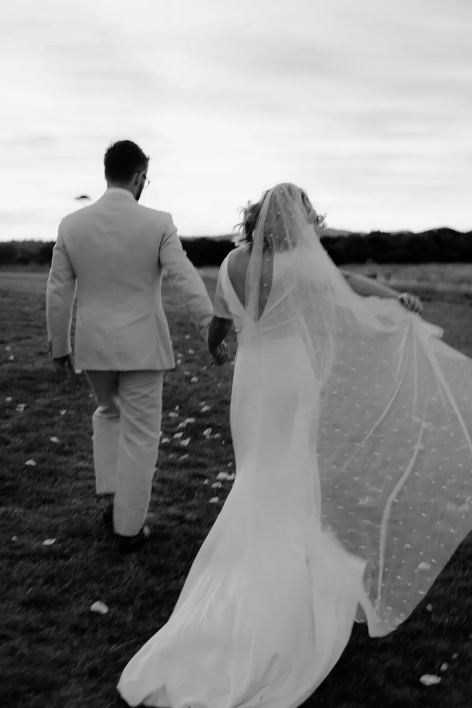 Black and white photograph of Jemmah and Jack walking together with her veil behind them, at Thalia Haven on the East Coast of Tasmania, Australia, for an Elopement Collective elopement. Photograph by Joey Willis.