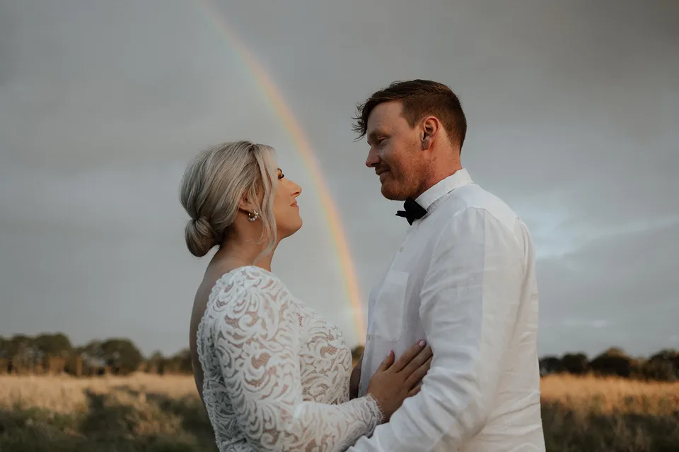 Matt and Laura looking at each other beneath a rainbow over the paddocks, in the Tweed Valley, NSW, for an Elopement Collective elopement. Photo by Bulb Creative.
