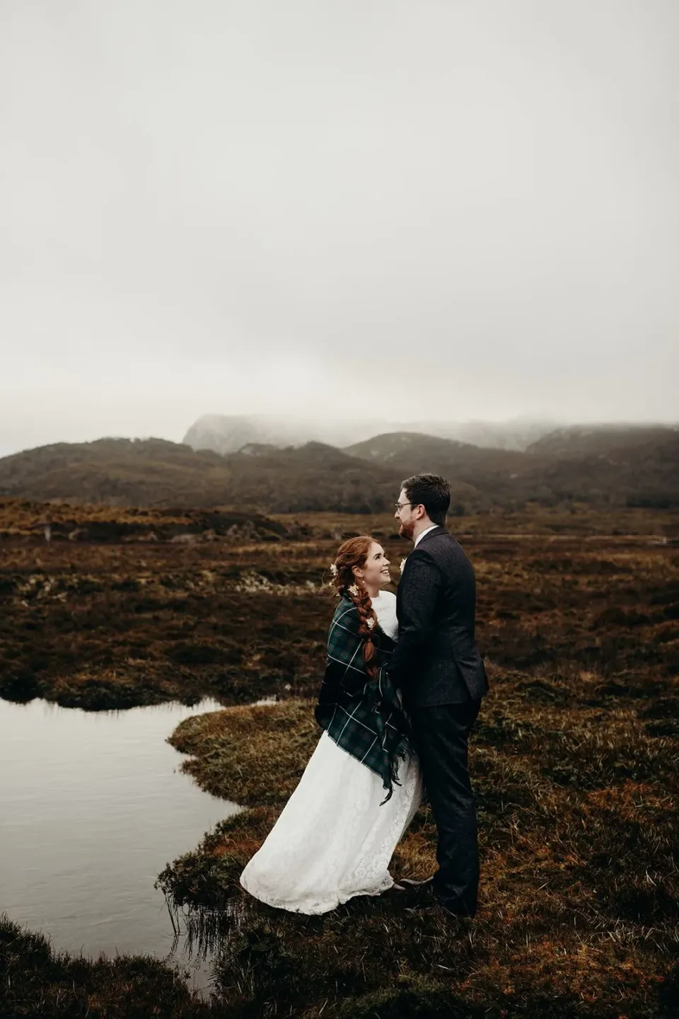 A couple embrace beside a still pool in a misty alpine landscape.