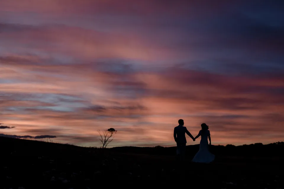 A wide view of the couple holding hands against the sunset sky, at Thalia Haven on the East Coast of Tasmania, Australia, for an Elopement Collective elopement. Photograph by Joey Willis.