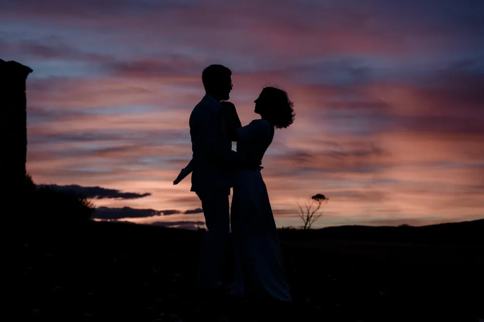 The couple embracing in silhouette beneath the glowing sunset, at Thalia Haven on the East Coast of Tasmania, Australia, for an Elopement Collective elopement. Photograph by Joey Willis.