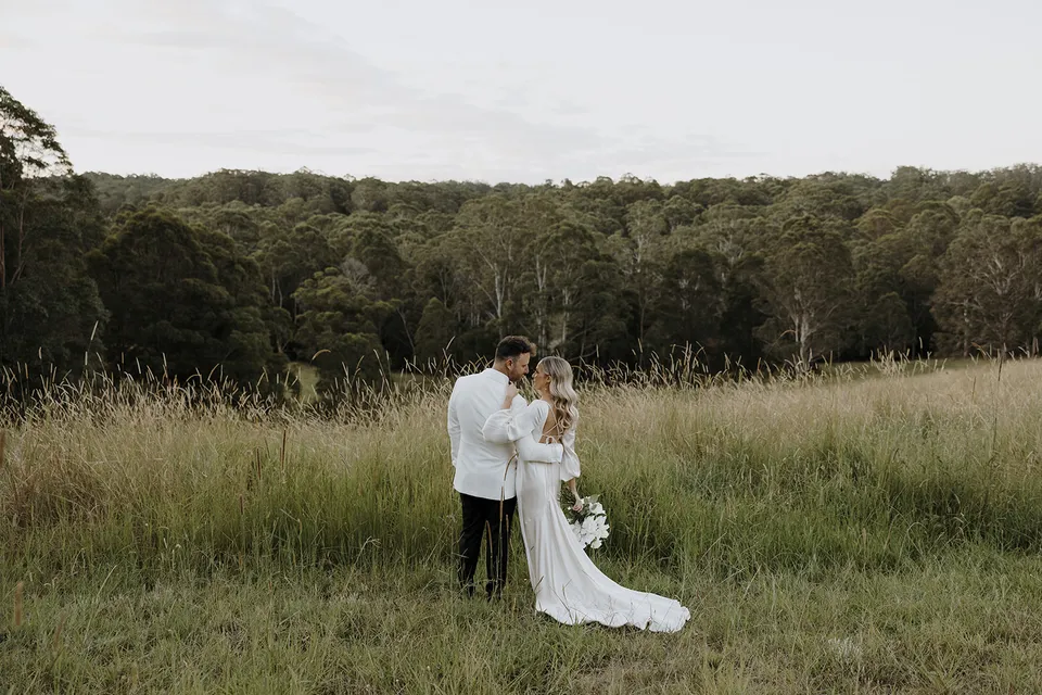 Gabrielle and Greg embracing in tall grass with a wooded ridge behind them at Roundhill Retreat in Obi Obi, Sunshine Coast Hinterland, for their Elopement Collective elopement.