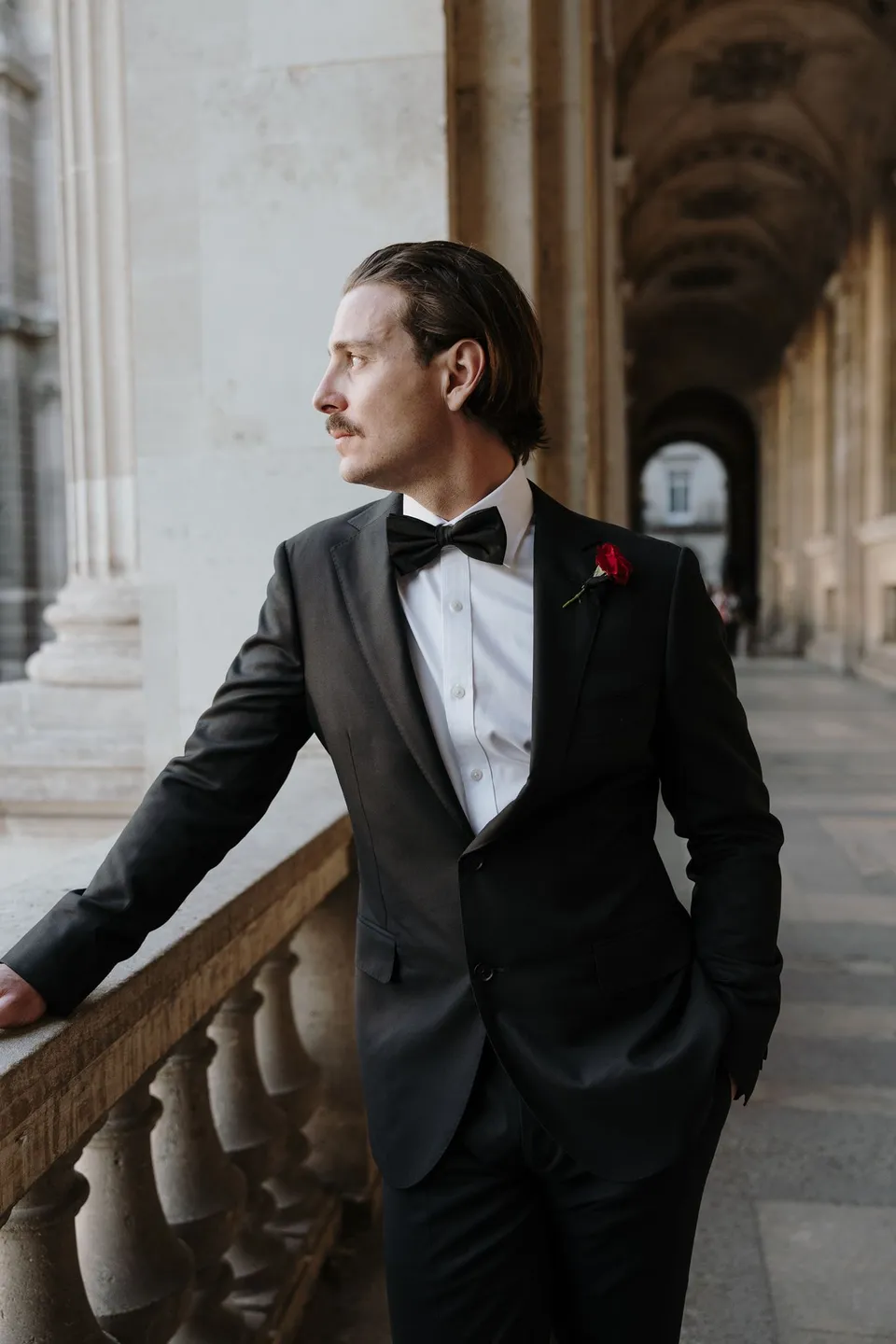 Samuel leaning against a stone balustrade beneath the Louvre’s arches in a black tuxedo with a red buttonhole by Ivy Bleu, at the Louvre in Paris, France, during an Elopement Collective elopement. Photograph by George Bowden.