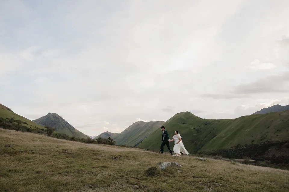 Jaeda and Adrian walking together across the grassy hillside near Moke Lake beneath a wide evening sky, at Moke Lake, Queenstown, New Zealand, for an Elopement Collective Queenstown elopement. Photograph by Bulb Creative.