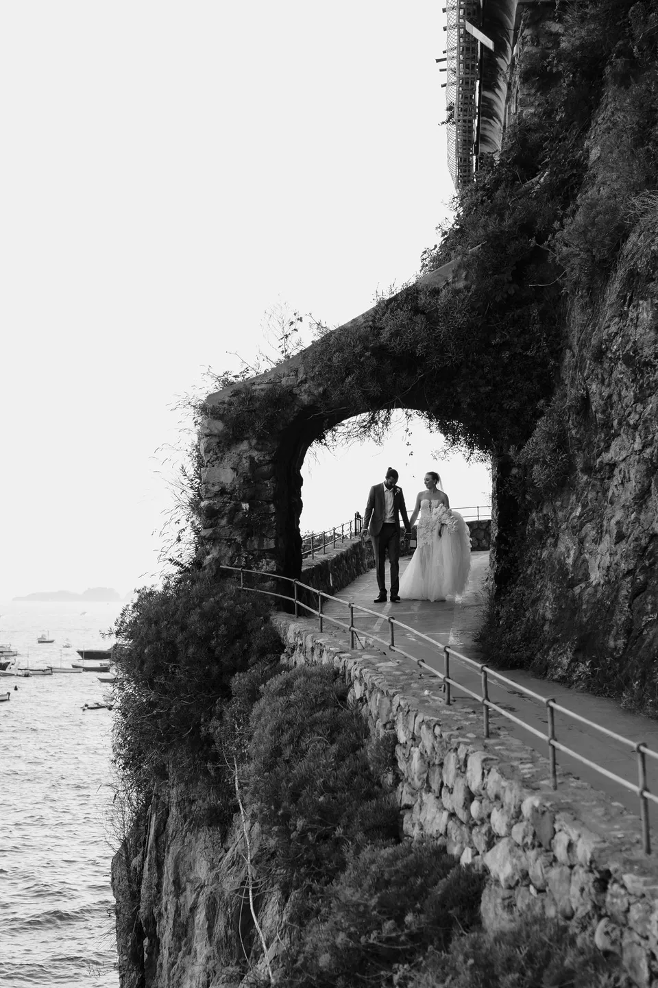 Black and white view of Skyler and Zach standing beneath an ivy-covered stone arch on a path above the sea, in Positano on the Amalfi Coast, Italy, for an Elopement Collective elopement. Photograph by Jason Corroto.