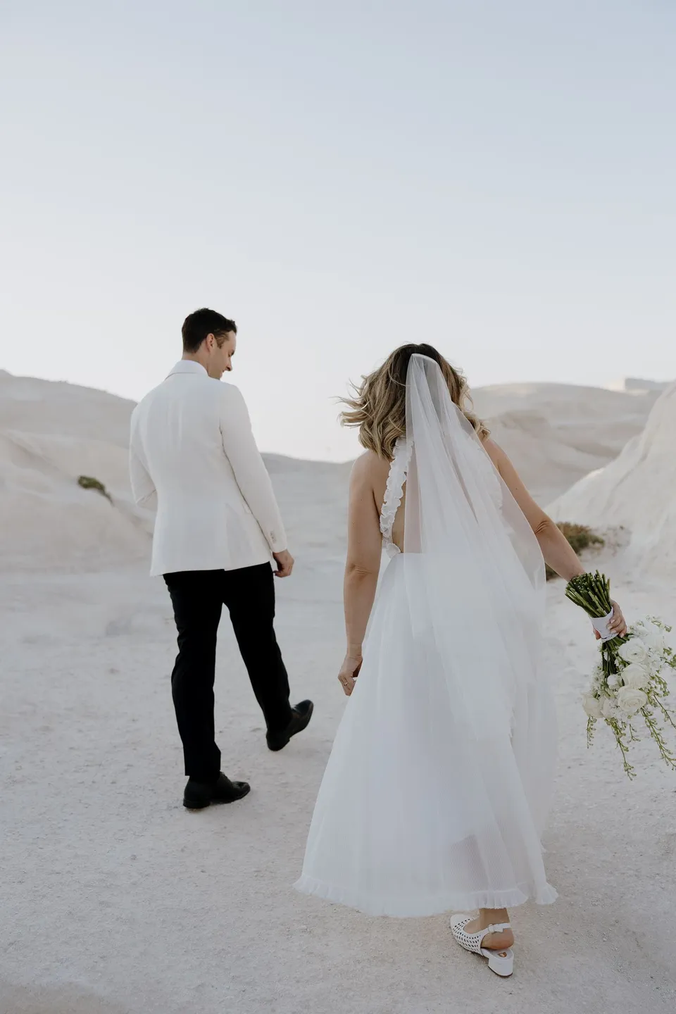 Rear view of Georgina and Brad holding hands as they walk across white rocks, with her veil and Giasemi bouquet trailing behind, at Sarakiniko on Milos in the Cyclades, Greece, during an Elopement Collective elopement. Photograph by George Bowden.