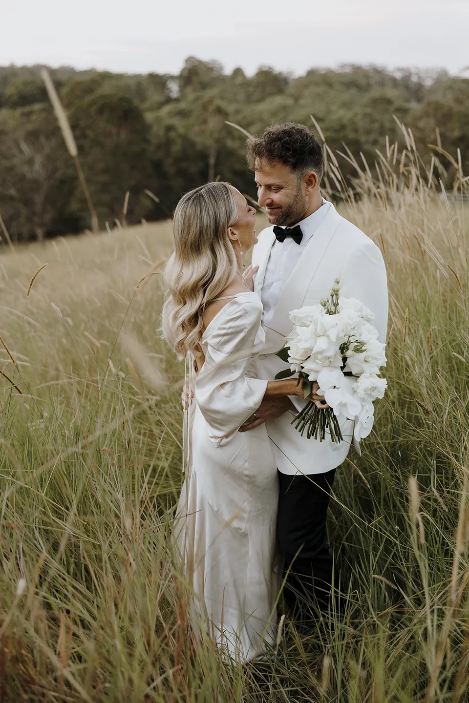Gabrielle holding her white bouquet as Greg smiles at her among the tall paddock grasses at Roundhill Retreat in Obi Obi, Sunshine Coast Hinterland, for their Elopement Collective elopement.