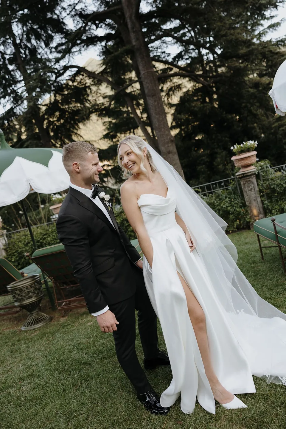 Zoe and Charlton smiling at each other on a garden path beneath tall trees, with her veil drifting to one side, at Passalacqua in Moltrasio, Lake Como, Italy, for an Elopement Collective elopement. Photo by George Bowden.