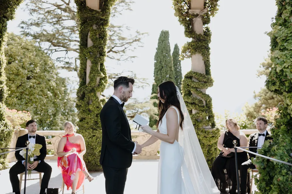 Lyndal reading from a vow book while holding Lachlan’s hand, with guests seated close by beneath Villa del Balbianello’s ivy-covered loggia, at Lake Como in Lombardy, Italy, for an Elopement Collective elopement. Photograph by George Bowden.
