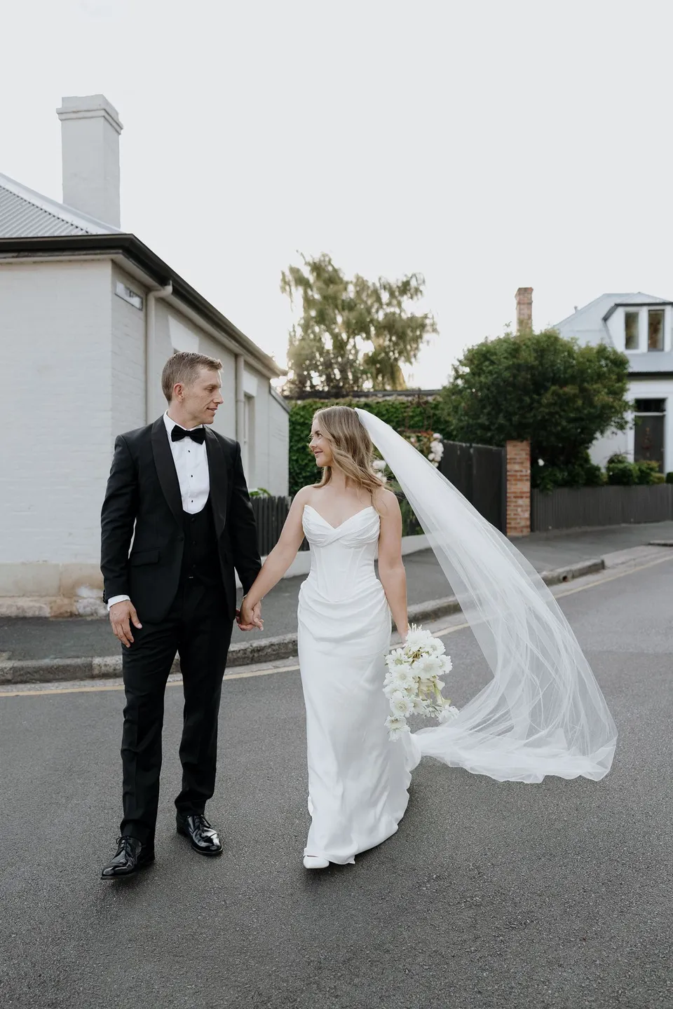 Tanesha and Andrew walking hand in hand past white cottages, with her veil lifting and her bouquet by The Romantics in hand, at Battery Point in Hobart, Tasmania, Australia, for an Elopement Collective summer elopement. Photograph by George Bowden.