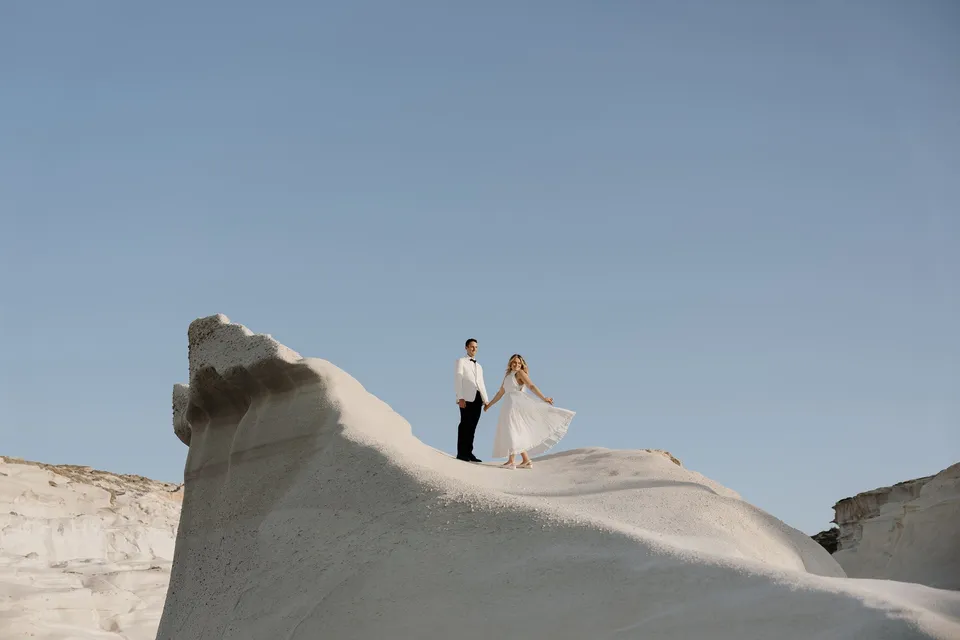Georgina and Brad walking along a sculpted white rock ridge beneath a pale sky, with her dress lifted above the ground, at Sarakiniko on Milos in the Cyclades, Greece, during an Elopement Collective elopement. Photograph by George Bowden.