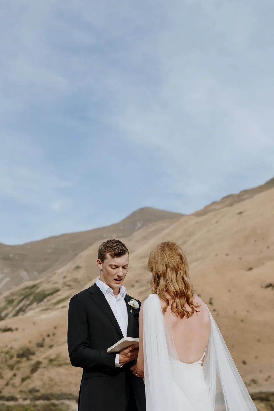 Hamish reading his vows to Neve beneath an open sky, with golden hills behind them, at Moke Lake in Queenstown, New Zealand, for an Elopement Collective Queenstown elopement. Photo by George Bowden.