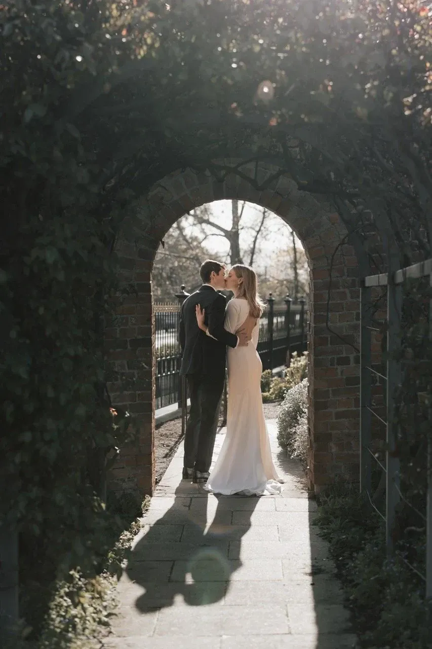 A couple embrace beneath a stone garden arch in dappled sunlight.