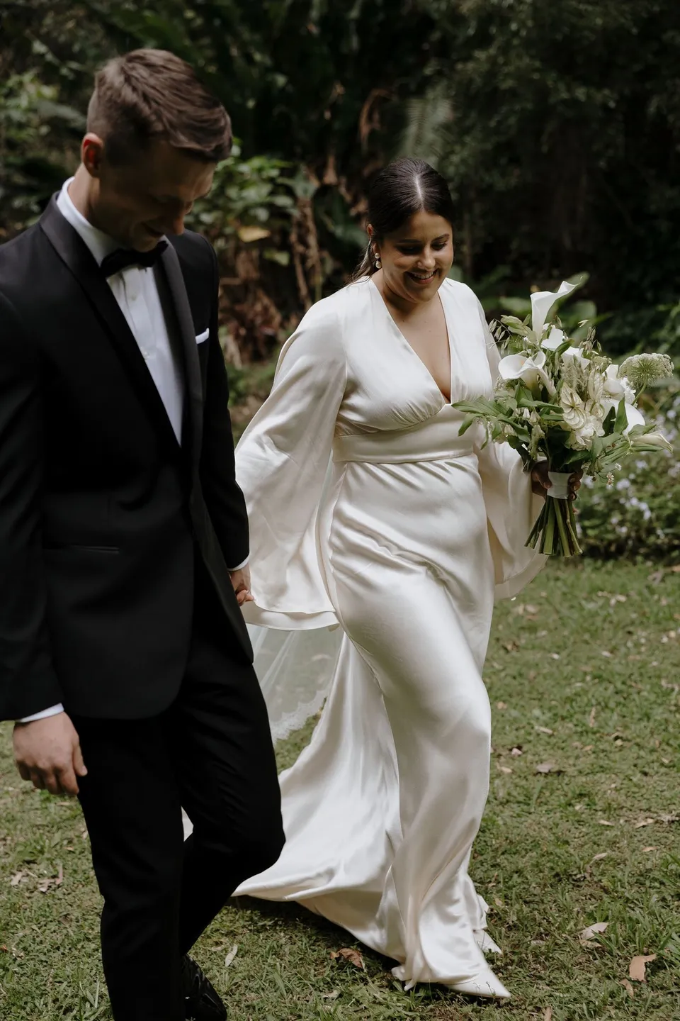 Laura and Matthew walking across the lawn with her white and green bouquet. At Fig Tree Villa in Ewingsdale, Byron Bay, for an Elopement Collective elopement. Photograph by George Bowden.