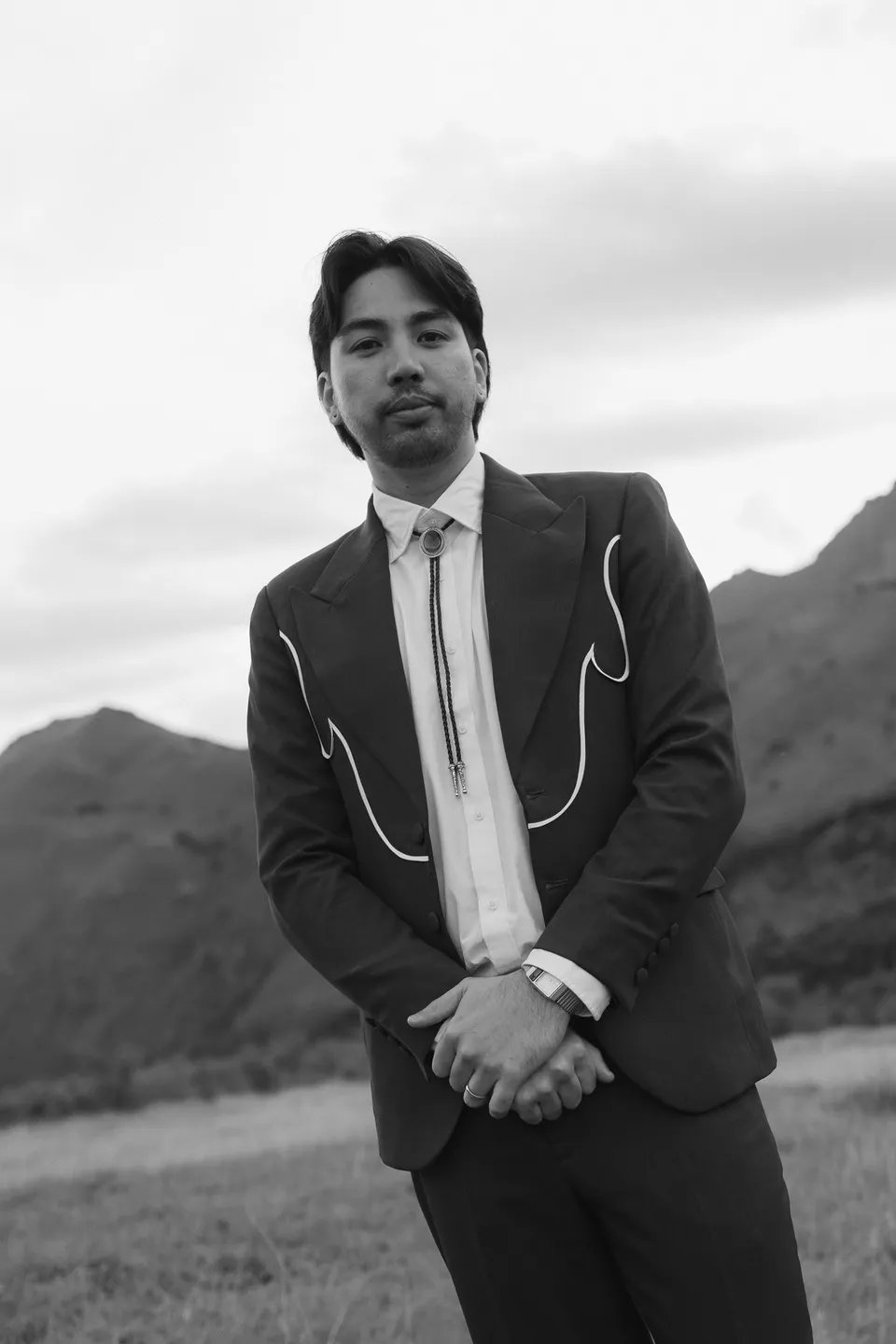 A black and white portrait of Adrian in his piped suit and bolo tie, with the hills behind him, at Moke Lake, Queenstown, New Zealand, for an Elopement Collective Queenstown elopement. Photograph by Bulb Creative.