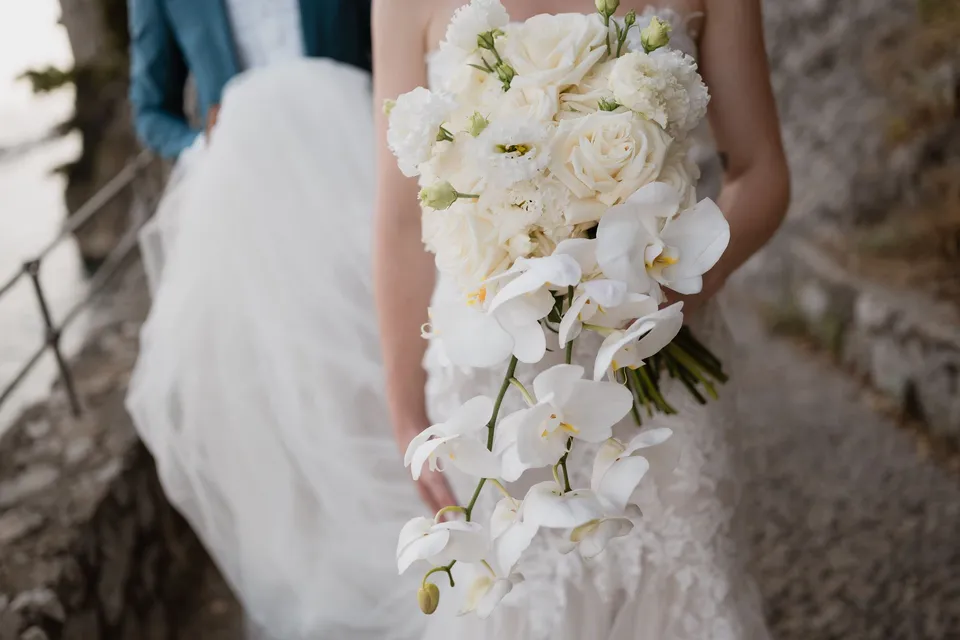 Close view of Skyler’s white rose and cascading orchid bouquet by Flora Garden - Positano against the lace of her wedding dress, in Positano on the Amalfi Coast, Italy, for an Elopement Collective elopement. Photograph by Jason Corroto.