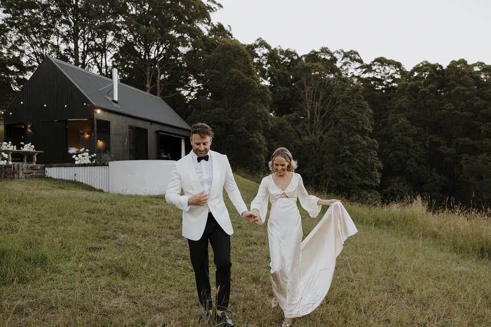 Gabrielle and Greg walking hand in hand through the paddock with the dark cabin and festoon lights behind them at Roundhill Retreat in Obi Obi, Sunshine Coast Hinterland, for their Elopement Collective elopement.