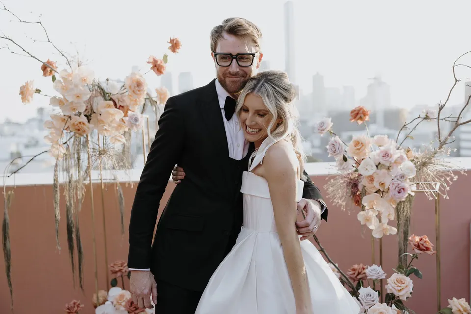 Adam embracing Danielle from behind as she smiles among the terrace flower arrangements, during Danielle and Adam’s Elopement Collective elopement at The Calile Hotel in Fortitude Valley, Brisbane.