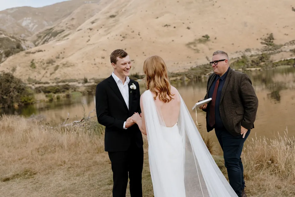 Hamish laughing with Neve and celebrant Josh Withers during their outdoor ceremony, at Moke Lake in Queenstown, New Zealand, for an Elopement Collective Queenstown elopement. Photo by George Bowden.