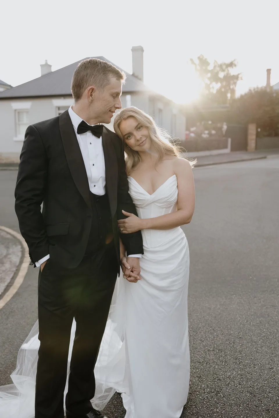 Tanesha resting her head against Andrew’s shoulder as they stand together in a sunlit cottage-lined street, at Battery Point in Hobart, Tasmania, Australia, for an Elopement Collective summer elopement. Photograph by George Bowden.