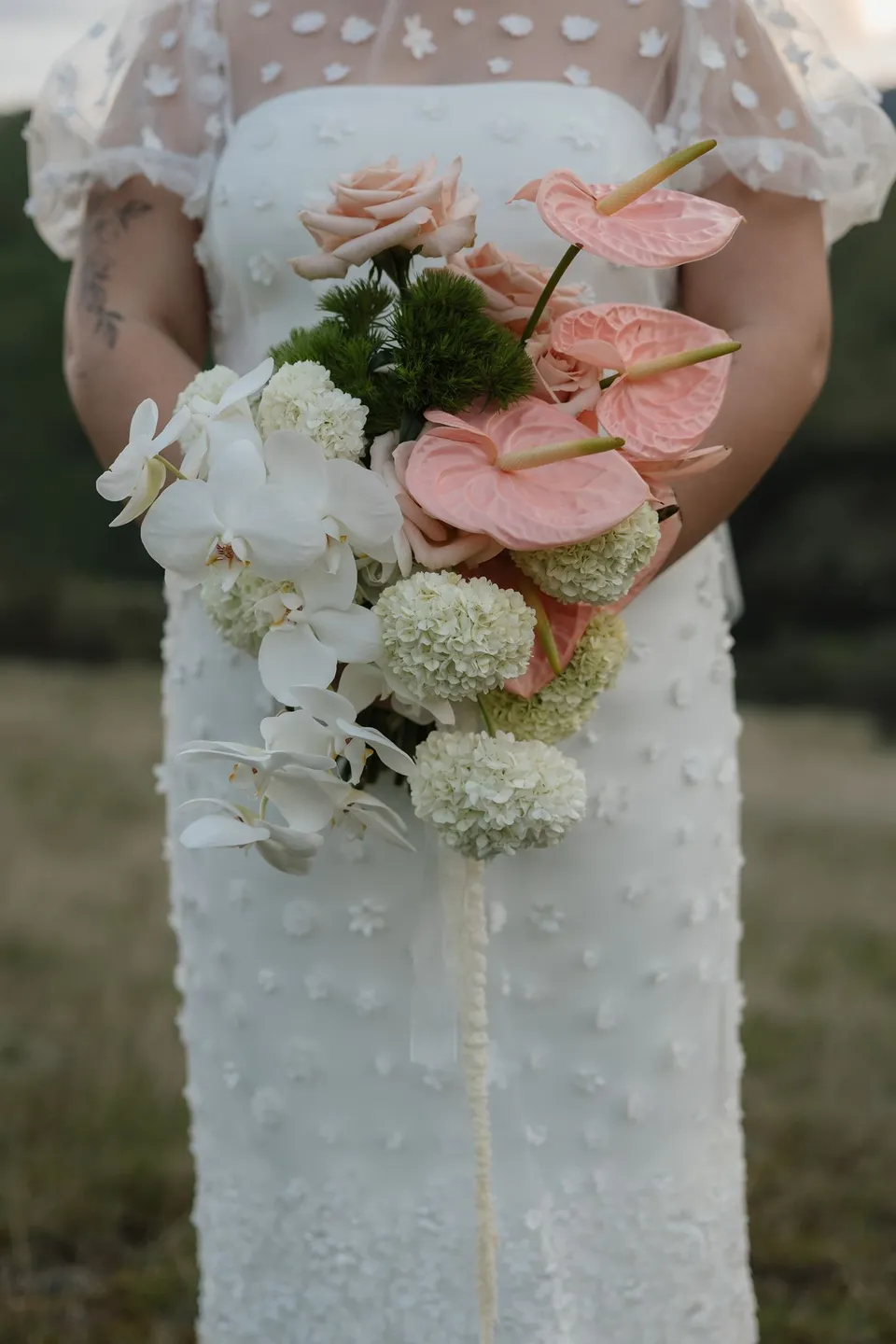 A close view of Jaeda’s Orange Blossom Designs bouquet of pink anthuriums, pale roses, white orchids and rounded white blooms against her daisy-appliqué dress, at Moke Lake, Queenstown, New Zealand, for an Elopement Collective Queenstown elopement. Photograph by Bulb Creative.