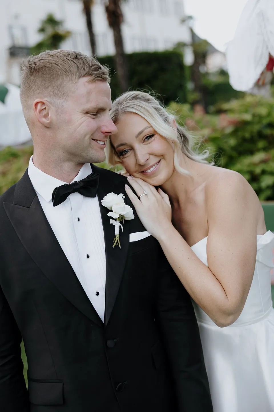 Zoe resting her cheek against Charlton and smiling, showing her softly waved hair and makeup by Valentina of Sunshine Brides and his white boutonniere by Garden of Love, at Passalacqua in Moltrasio, Lake Como, Italy, for an Elopement Collective elopement. Photo by George Bowden.