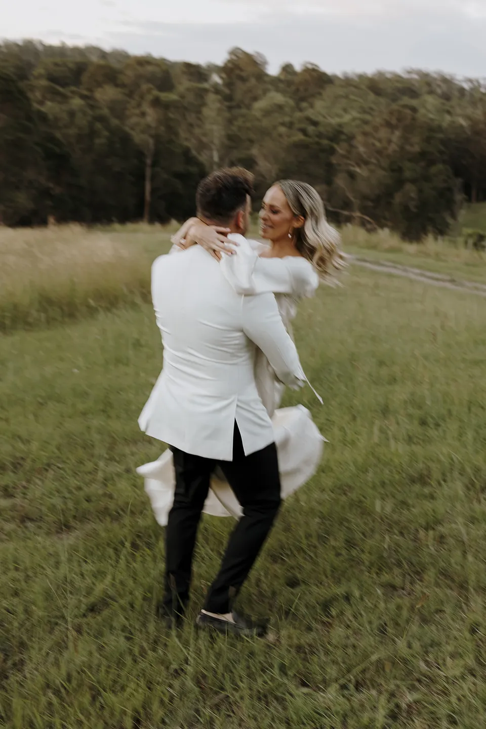 Greg carrying Gabrielle through the grassy paddock as she wraps her arms around his shoulders at Roundhill Retreat in Obi Obi, Sunshine Coast Hinterland, for their Elopement Collective elopement.