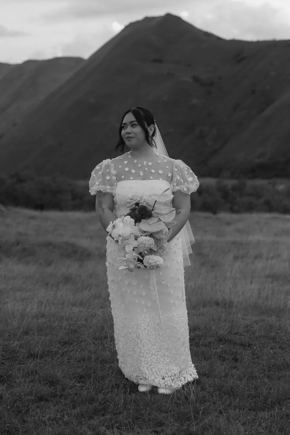 A black and white bridal portrait of Jaeda holding her Orange Blossom Designs bouquet beside the mountain landscape, at Moke Lake, Queenstown, New Zealand, for an Elopement Collective Queenstown elopement. Photograph by Bulb Creative.
