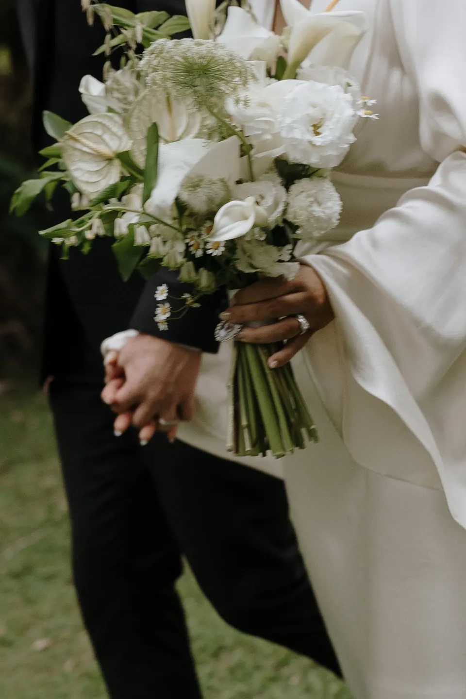Close-up of Laura's bouquet by The French Petal beside the couple's joined hands. At Fig Tree Villa in Ewingsdale, Byron Bay, for an Elopement Collective elopement. Photograph by George Bowden.