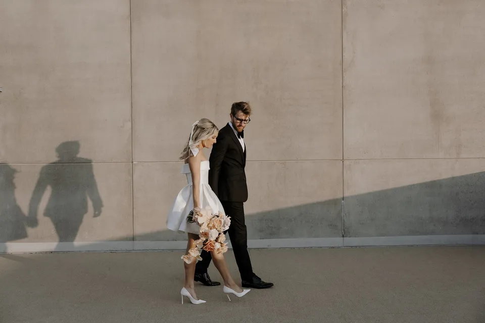 Danielle and Adam walking beside a plain rooftop wall with their shadows falling behind them, during Danielle and Adam’s Elopement Collective elopement at The Calile Hotel in Fortitude Valley, Brisbane.