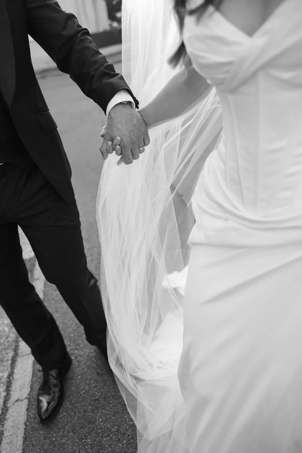 Black and white close-up of Tanesha and Andrew’s joined hands beside her draped white wedding dress, at Battery Point in Hobart, Tasmania, Australia, for an Elopement Collective summer elopement. Photograph by George Bowden.