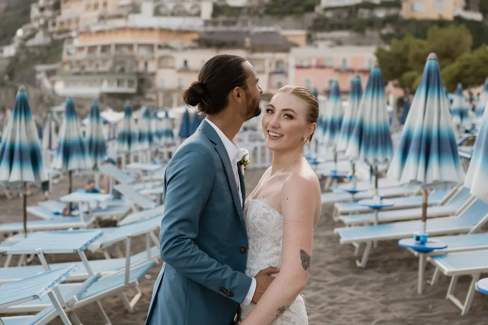 Zach kissing Skyler’s cheek as she smiles beside rows of blue beach umbrellas and loungers, in Positano on the Amalfi Coast, Italy, for an Elopement Collective elopement. Photograph by Jason Corroto.