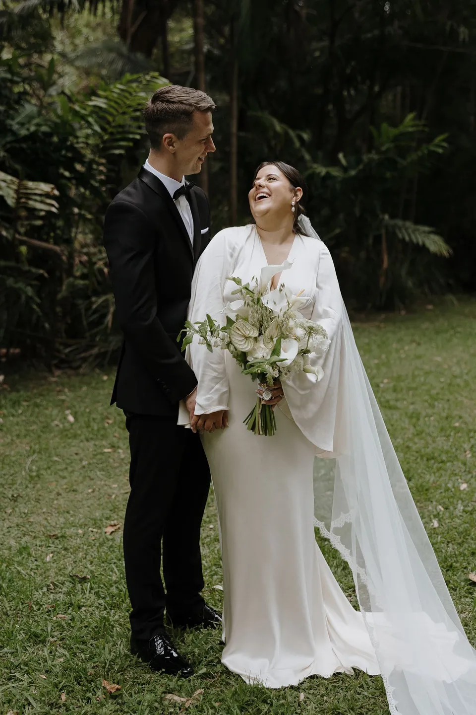 Laura and Matthew holding hands and smiling at each other on the lawn. At Fig Tree Villa in Ewingsdale, Byron Bay, for an Elopement Collective elopement. Photograph by George Bowden.