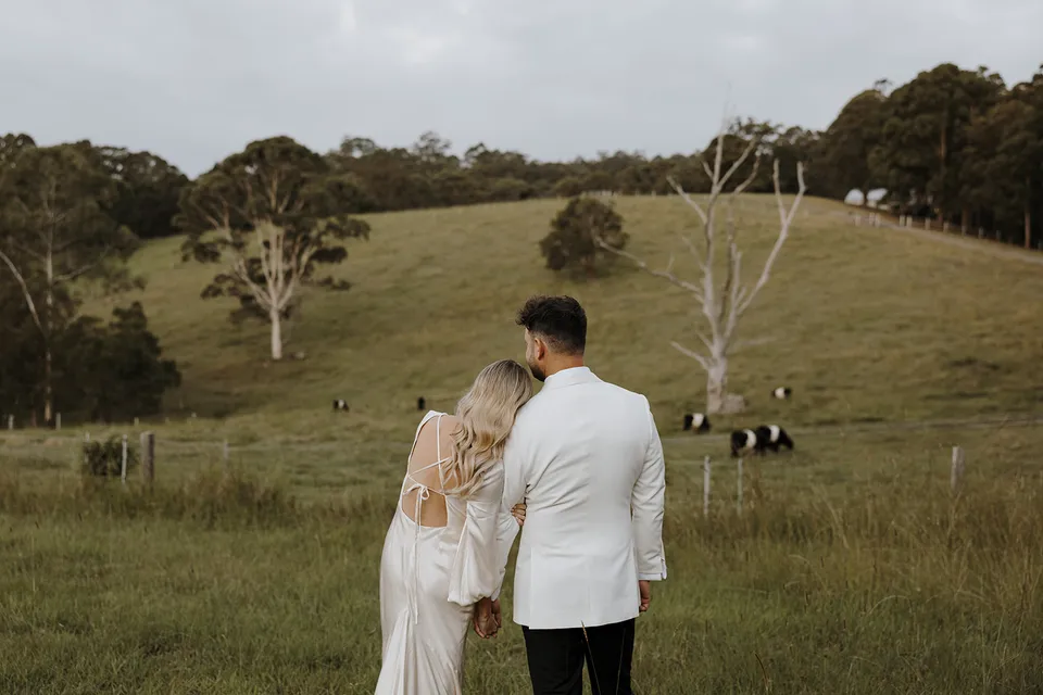 Gabrielle leaning on Greg’s shoulder as they look over rolling green paddocks and grazing cattle at Roundhill Retreat in Obi Obi, Sunshine Coast Hinterland, for their Elopement Collective elopement.