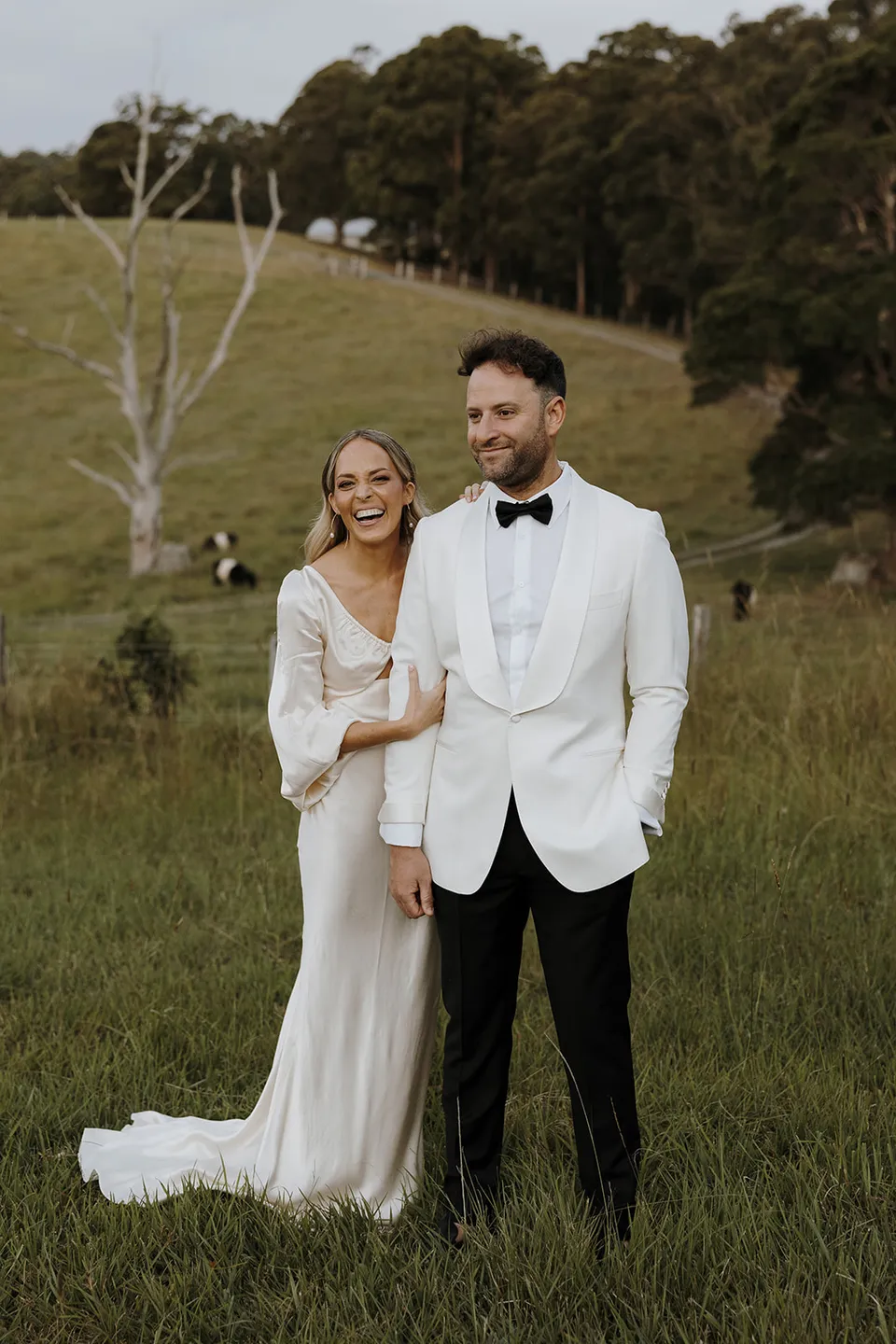 Gabrielle and Greg laughing together in a paddock with green hills and trees behind them at Roundhill Retreat in Obi Obi, Sunshine Coast Hinterland, for their Elopement Collective elopement.