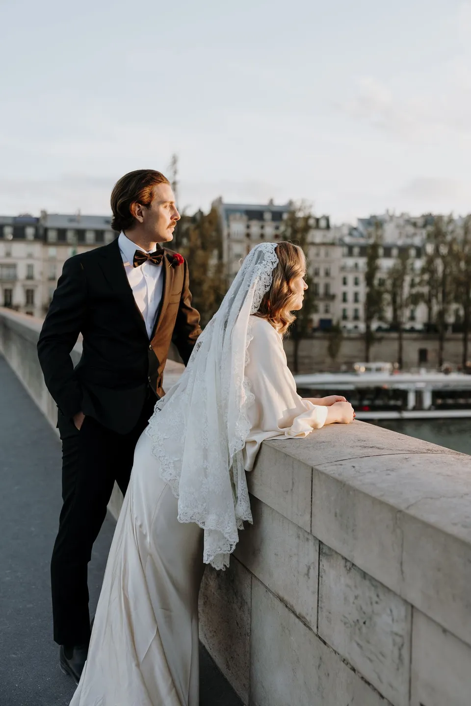 Kara and Samuel standing close beside a stone bridge balustrade above the Seine, with Parisian buildings across the river, beside the Seine in Paris, France, during an Elopement Collective elopement. Photograph by George Bowden.