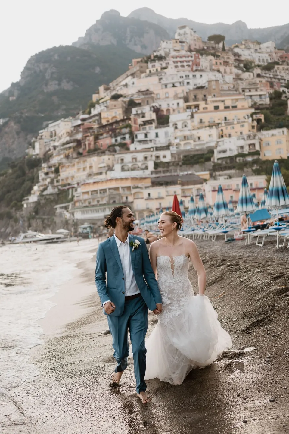 Skyler and Zach holding hands as they walk along the water’s edge, beneath the houses and mountains of Positano, in Positano on the Amalfi Coast, Italy, for an Elopement Collective elopement. Photograph by Jason Corroto.