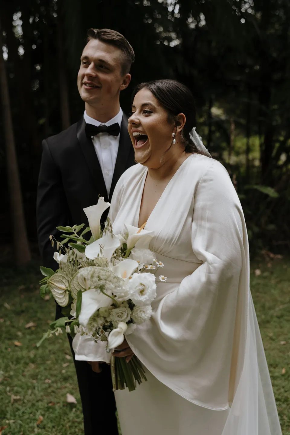 Laura laughing beside Matthew in the green garden. At Fig Tree Villa in Ewingsdale, Byron Bay, for an Elopement Collective elopement. Photograph by George Bowden.