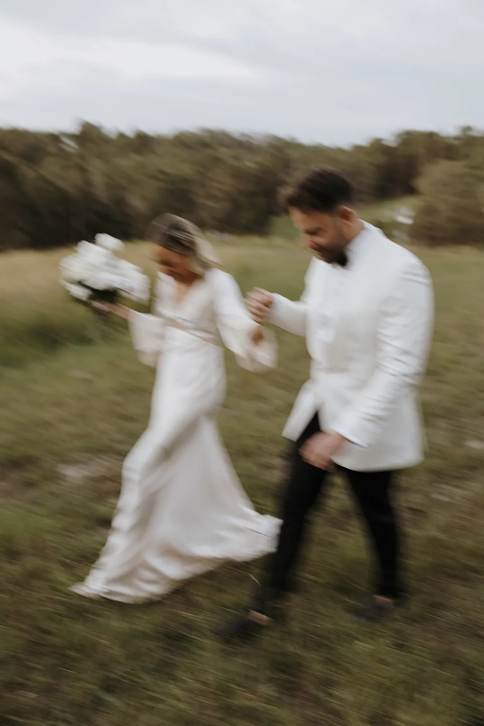 Softly blurred photograph of Gabrielle and Greg walking through the grass, holding hands and her white bouquet at Roundhill Retreat in Obi Obi, Sunshine Coast Hinterland, for their Elopement Collective elopement.