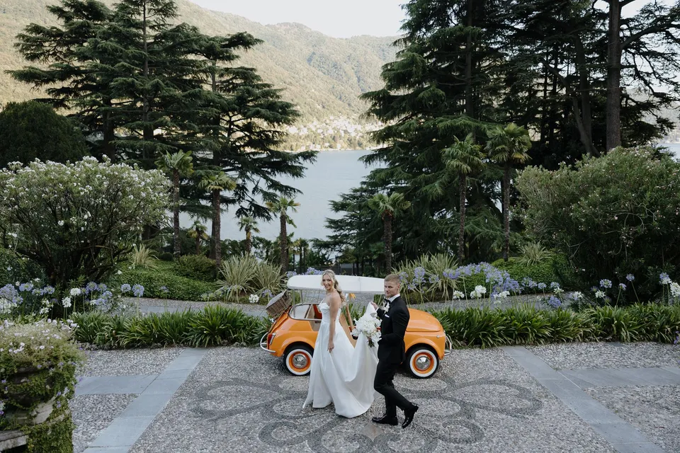 Wide view of Zoe and Charlton walking hand in hand across the cobbled driveway beside an orange car, with terraced gardens and the lake beyond, at Passalacqua in Moltrasio, Lake Como, Italy, for an Elopement Collective elopement. Photo by George Bowden.
