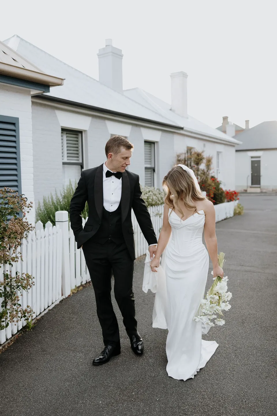 Tanesha and Andrew walking beside cottage gardens, looking down while she carries her white bouquet by The Romantics, at Battery Point in Hobart, Tasmania, Australia, for an Elopement Collective summer elopement. Photograph by George Bowden.
