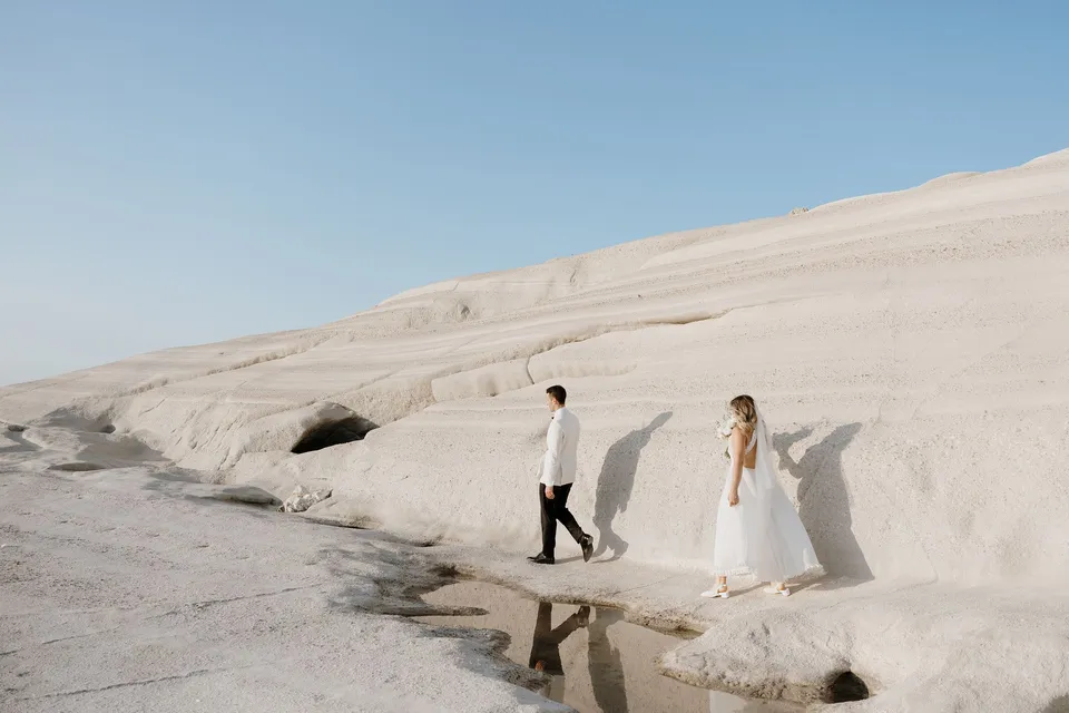 Georgina and Brad walking across pale rocks beside a small pool reflecting their figures, at Sarakiniko on Milos in the Cyclades, Greece, during an Elopement Collective elopement. Photograph by George Bowden.