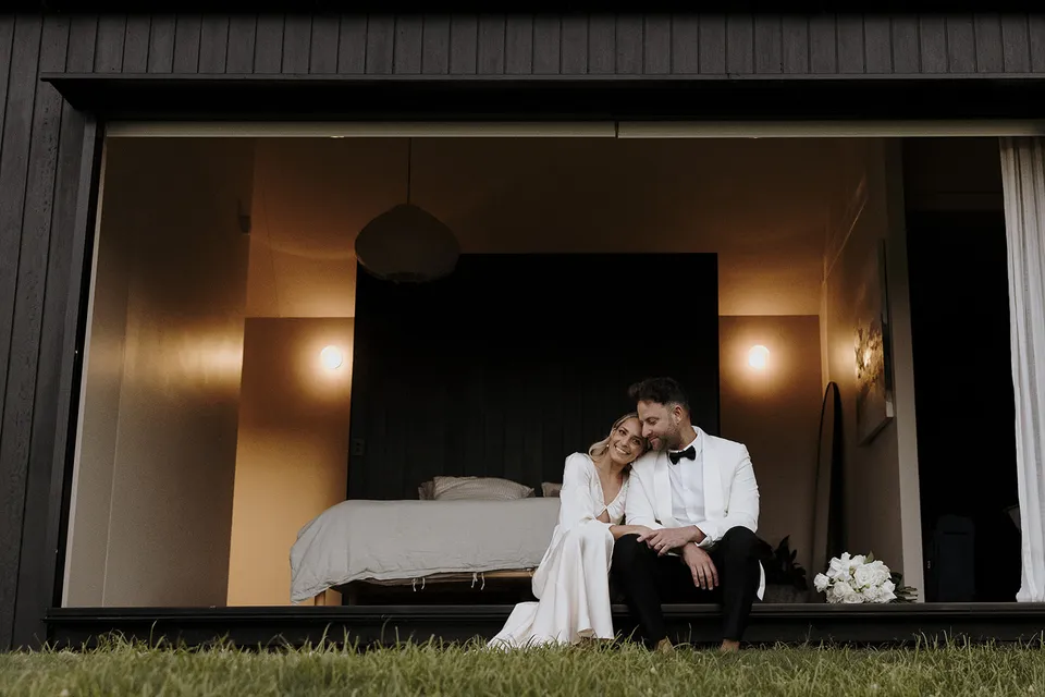 Gabrielle and Greg sitting together in the open bedroom doorway, framed by the cabin’s dark timber exterior at Roundhill Retreat in Obi Obi, Sunshine Coast Hinterland, for their Elopement Collective elopement.